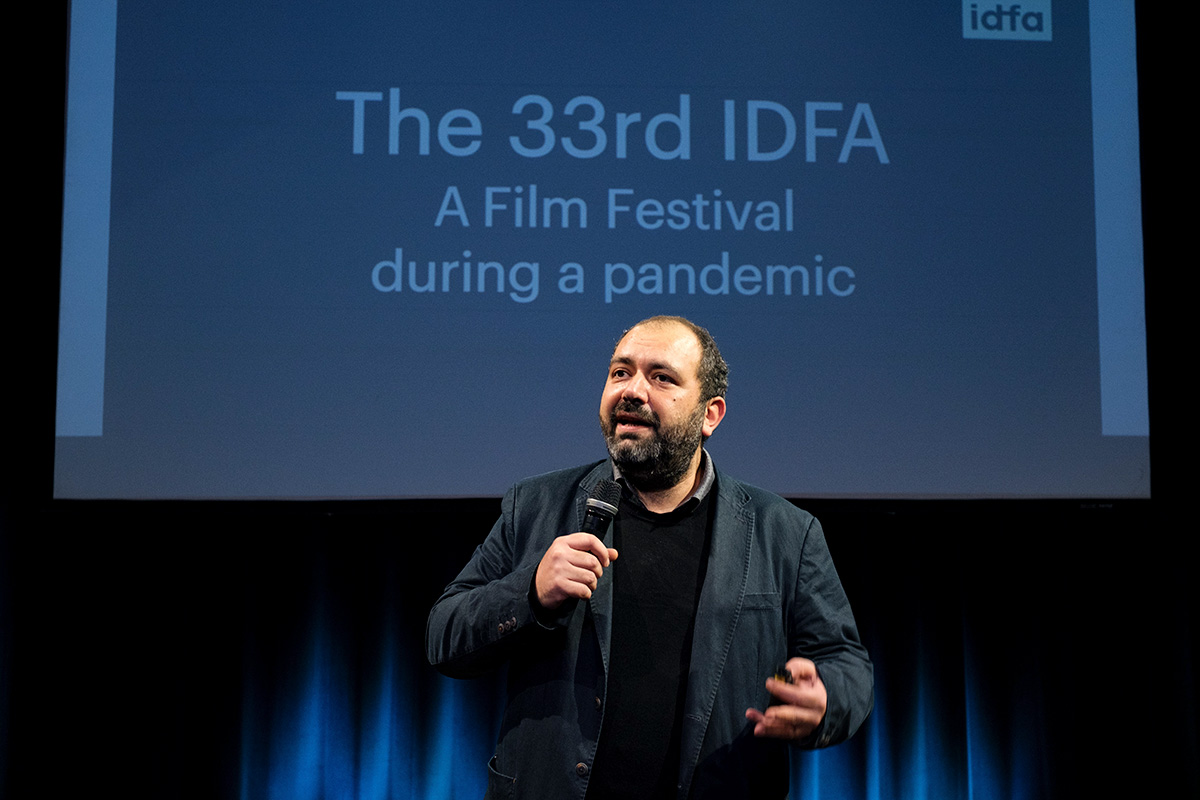 IDFA Artistic Director Orwa Nyrabia is a Syrian man with a beard and short hair. He is wearing a black jacket and a sweater and is addressing the in-person and virtual audience at IDFA 2020. There is a screen behind him. Photo: Coen Dijkstra. Courtesy of IDFA