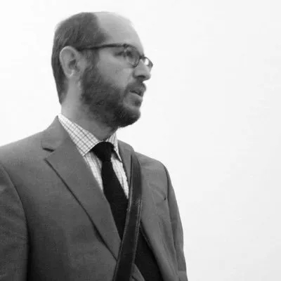Black and white headshot of a middle aged male with a short beard and glasses wearing a suit and looking off to the right